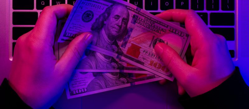 Dollar bills in male hands on the background of a laptop keyboard in neon lighting, top view.